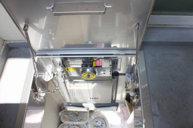 Metalik endüstriyel makine, vites kolları, düğmeler ve uyarı etiketleri içeriyor.