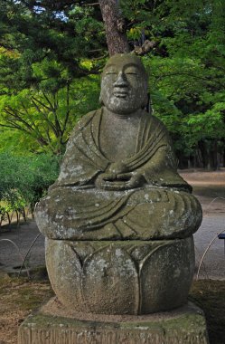 Yıpranmış taş Buda heykeli yemyeşil ağaçların arasında duruyor.