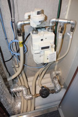 Beyaz gaz sayacı izole borular ve elektrik kablolarıyla çevrili.