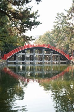 Yeşil ağaçlar arasında sakin suyun üzerine yansıyan kırmızı kemerli köprü.