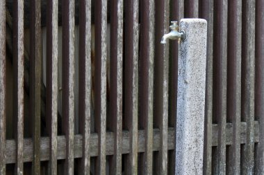 Eski metal su musluğu ahşap çitin yanındaki beton direğe bağlanmış.