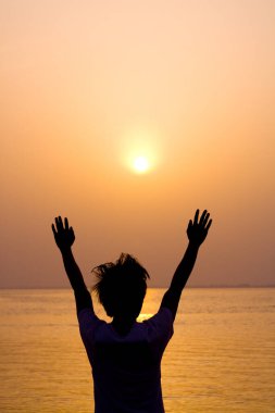 Gün batımında kumsalda kollarını açmış genç bir kadının silueti. 