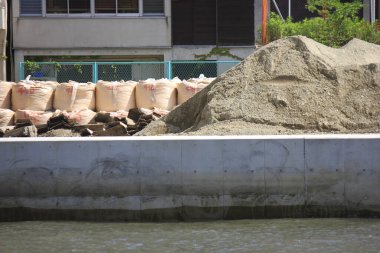 Dışarıdaki beton duvarın yanında istiflenmiş çuvalların yanında büyük kum yığını var.