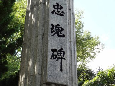 Yeşil ağaçlar arasında Çince karakterlerin yer aldığı uzun taş heykel.