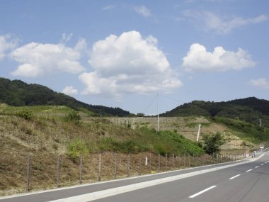 Boş kırsal yol mavi gökyüzünün altında çimenli tepelerin yanında kıvrılıyor.