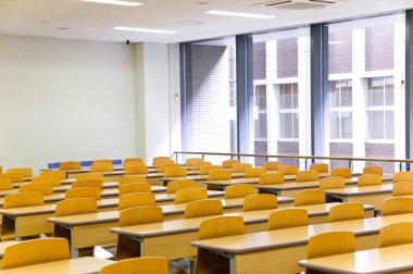 Büyük pencereli boş okul odası, boş ahşap masalar ve sandalyeler.