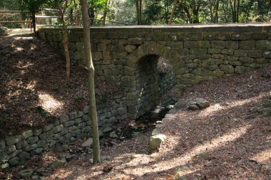 Ağaçlarla çevrili küçük bir dere üzerinde taş köprü.