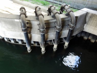 Suyun üstündeki beton duvardan geçen büyük endüstriyel borular.