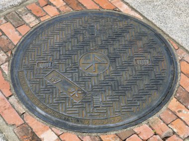 Kırmızı tuğlaların arasında duran yuvarlak metal rögar kapağı.