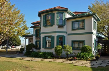 Yeşil kepenkleri ve manikürlü çalıları olan iki katlı bir ev.