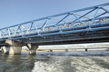 Mavi çelik köprü, temiz gökyüzünün altında geniş bir nehri kaplıyor.