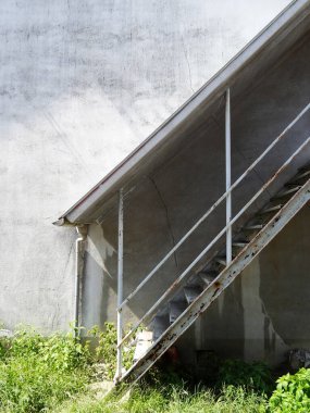 Güneş ışığı yıpranmış beton duvarın yanındaki metal merdivenlere çarpıyor.