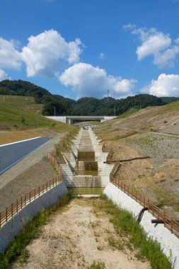 Beton drenaj kanalı mavi gökyüzünün altındaki çimenli tepelerde akıyor.