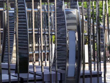 Açık havada dikey parmaklıkların arasında duran yansıtıcı metal heykeller.