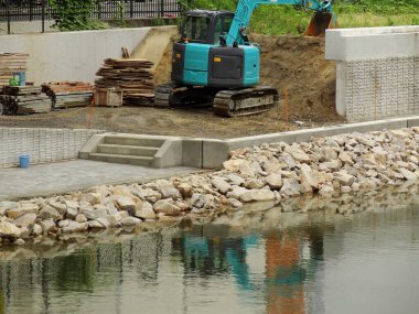 Mavi kazıcı beton nehir setinin yakınındaki toprağı kazıyor.