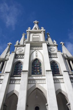 Beyaz taş kilise cephesi, kuleler, sivri kemerler ve mavi gökyüzüne karşı boyalı camlar..