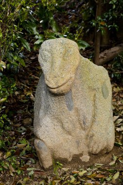 Yeşil yapraklı bitkilerin arasında duran yıpranmış taş hayvan heykeli.