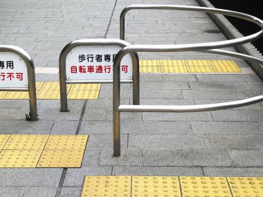 Sarı dokunsal asfalt çizgileri metal bariyerlerin yanındaki gri kaldırımı geçiyor