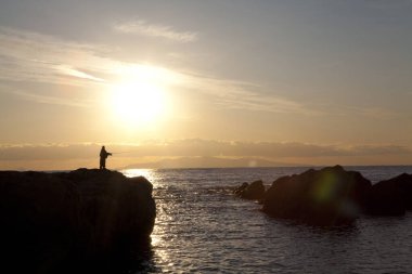 Gün batımında kayadan balık tutan insan silueti.