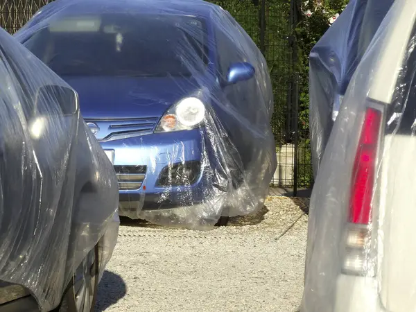 Açık havada park halindeki araçların arasında plastikle kaplı mavi bir araba.