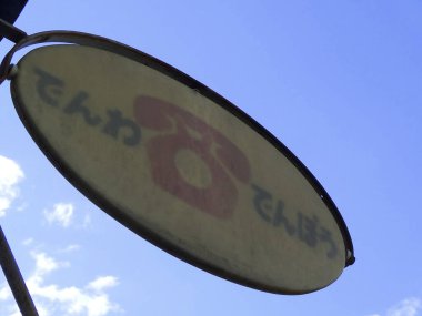 Üzerinde telefon sembolü ve Japon Hiragana metni olan mavi gökyüzüne asılı yıpranmış bir oval tabela Denwa 