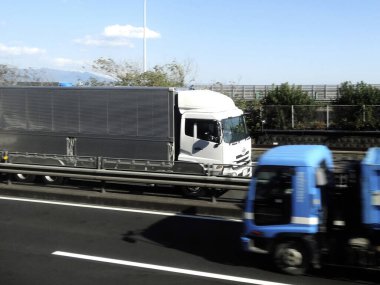 Otoyolda mavi aracın yanında giden büyük beyaz bir kamyon.