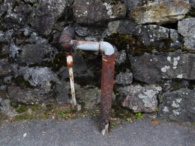 Eski taş duvarın karşısında duran paslı metal su borusu.