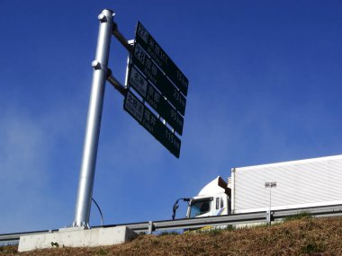 Büyük bir kamyon açık mavi gökyüzünün altında otoyol tabelasının yanından geçiyor.