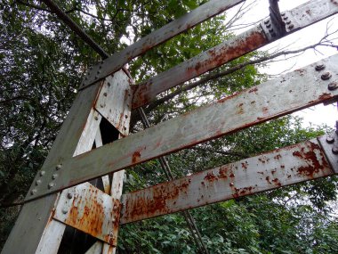 Paslı metal kirişler dışarıda yeşil yapraklı ağaçların arasında kesişiyor.