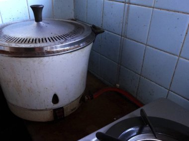 Eski pilav pişiricisi mavi fayanslı mutfak duvarının yanında oturuyor.