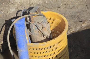 Sarı plastik sepet, açık havada mavi hortumun yanında çamurlu metal bir nesne tutuyor.