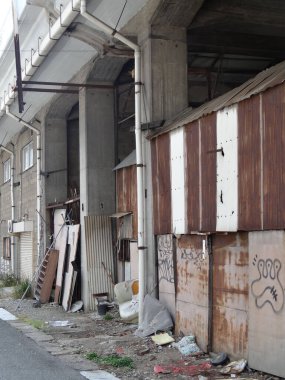 Terk edilmiş sanayi binasının dış tarafında paslanmış metal paneller, grafiti kalıntıları ve dağınık toprak parçaları görülüyor.