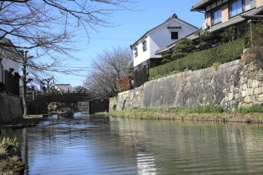 Geleneksel Japon evleri mavi gökyüzünün altındaki sakin nehir yolu boyunca yansıyor.