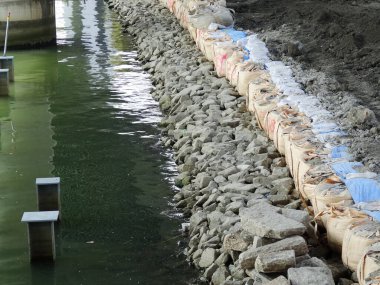 Kum torbaları ve inşaat malzemelerinin yer aldığı yeşil suyun yanındaki kayalık kıyı şeridi