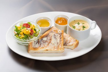 Kahvaltı tabağı, tost, salata ve iki kase çorba, beyaz tabakta servis edilir.