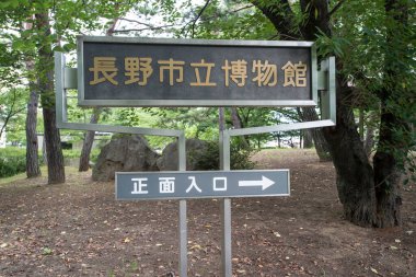 Nagano Şehir Müzesi ana girişi için imza, ağaçların ve bahçelerin arasına yerleştirilmiş, sağı işaret ediyor..