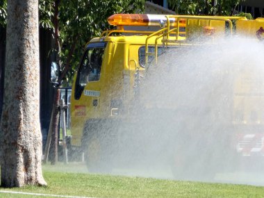 Sarı kamu kamyonu yeşil çimenli arazide su püskürtüyor.