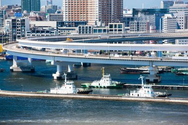 Gemiler yükseltilmiş otoyolların altındaki bir limanda seyrediyor. Arkasında yoğun bir şehir silüeti var..
