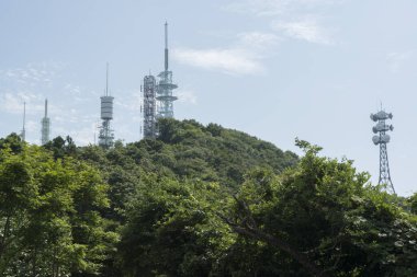 Çoklu telekomünikasyon kuleleri parlak bir gökyüzünün altındaki yoğun ormanlık yeşil tepeye hakim..