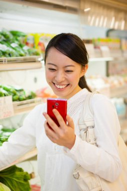 Markette akıllı telefon alışverişi yapan gülümseyen Japon kadın, telefondaki alışveriş listesini kontrol ediyor.