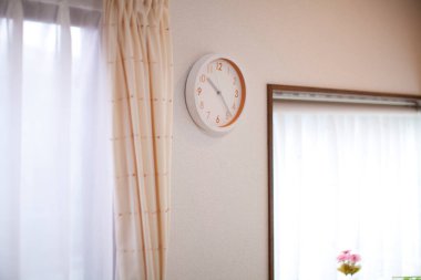 Pencerenin yanında asılı duvar saati, perdeler ve çiçeklerle