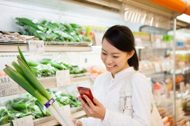 Markette akıllı telefon alışverişi yapan gülümseyen Japon kadın, telefondaki alışveriş listesini kontrol ediyor.