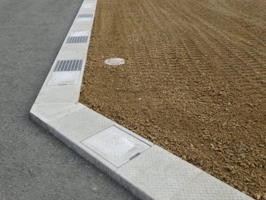Metal drenaj ızgaralarının olduğu beton kaldırımın yanında taze tırmıklanmış çakıllar.