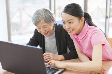 Genç Asyalı kadın yaşlı kadına evde dizüstü bilgisayar kullanmayı öğretiyor.  