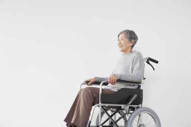 Beyaz arka planda tekerlekli sandalyede oturan gülümseyen yaşlı Asyalı kadın. 