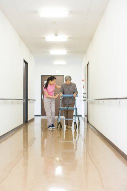 Koridorda yürüyen yaşlı bir kadına yardım eden kadın.
