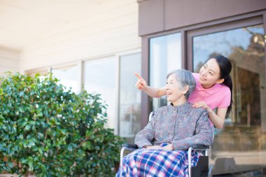 Japon kadın bakıcı, tekerlekli sandalyedeki yaşlı Asyalı kadına yardım ediyor.
