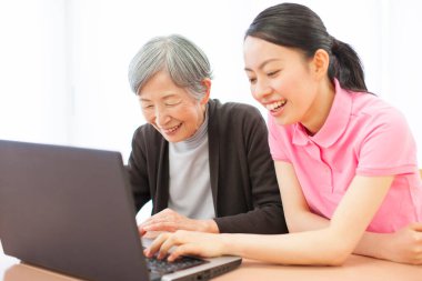 Genç Asyalı kadın yaşlı kadına evde dizüstü bilgisayar kullanmayı öğretiyor.  