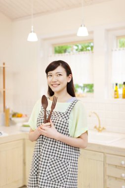Asyalı kadın mutfak aletlerini elinde tutuyor ve kameraya gülümsüyor.