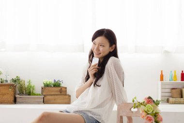 Güzel Asyalı genç bir kadının portresi. Açık ev iç mekanında akıllı telefon kullanıyor.